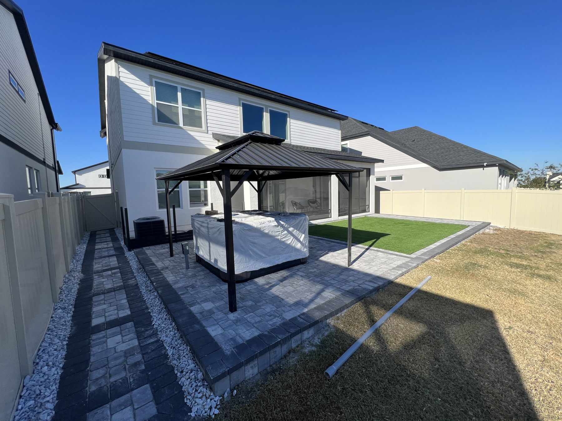 Finished grey paver patio with a dark hardtop pergola over a hot tub, artificial turf to one side and a white stone border along the fence line
